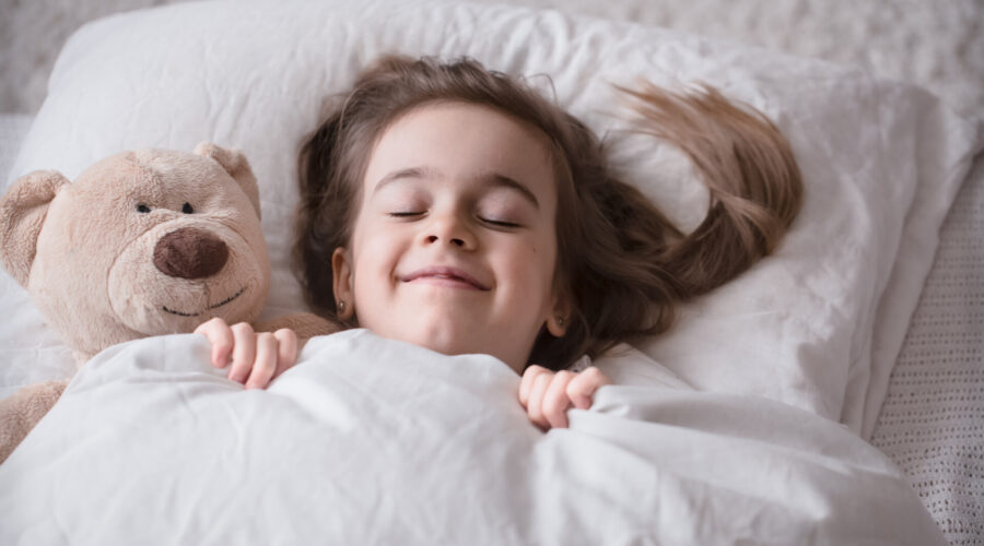 Cute little girl sleeps sweetly in a white cozy bed with a soft bear toy, the concept of children’s rest and sleep