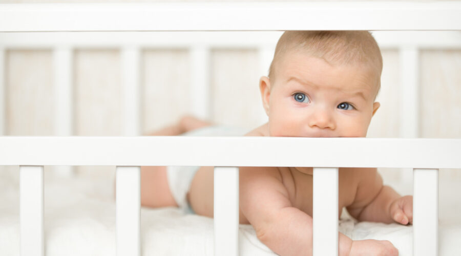Cute baby on a tummy in a white cot looking across the room through the wooden frame, itching gums, sucking the wooden board of the cot, teething proccess. Family, baby development concept photo